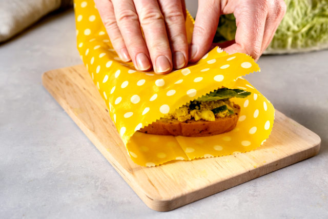 Zero waste and plastic free concept. Woman hands wrapping a healthy vegan sandwich in beeswax food wrap for lunch. Eco-friendly, reusable wax cloth and cotton bags