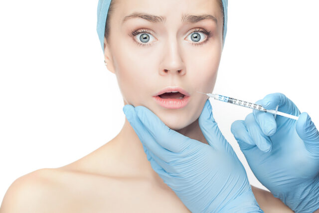 Attractive surprised and scared woman at plastic surgery with syringe in her face on white background