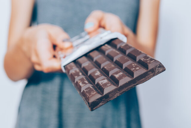 Female's hands holding unwrapped dark chocolate bar, close-up. Unrecognizable young woman standing and offering cocoa dessert.