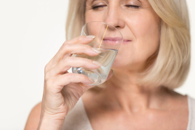 Healthy thirsty senior old woman drinking filtered pure fresh clean water, middle aged mature lady holding glass hydrating body isolated on white grey studio background, dehydration hydration concept